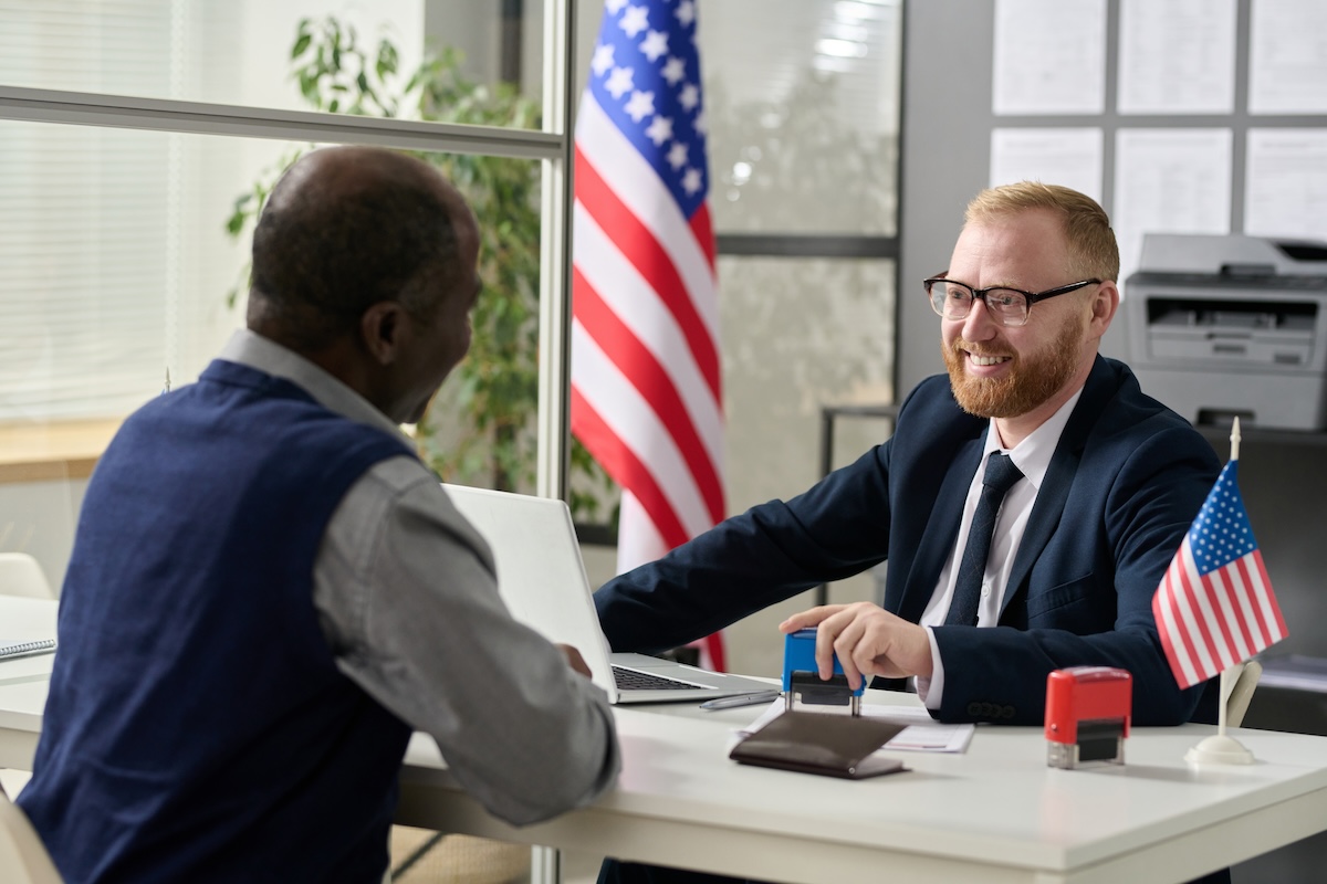 Dos personas conversan en un escritorio con una computadora, sellos y banderas de Estados Unidos