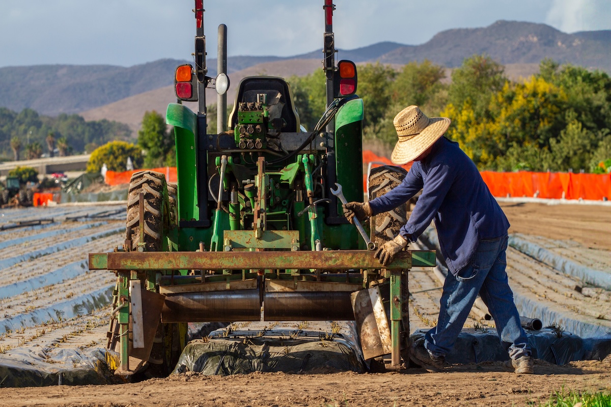 Persona con sombrero y guantes trabajando junto a un tractor en un campo